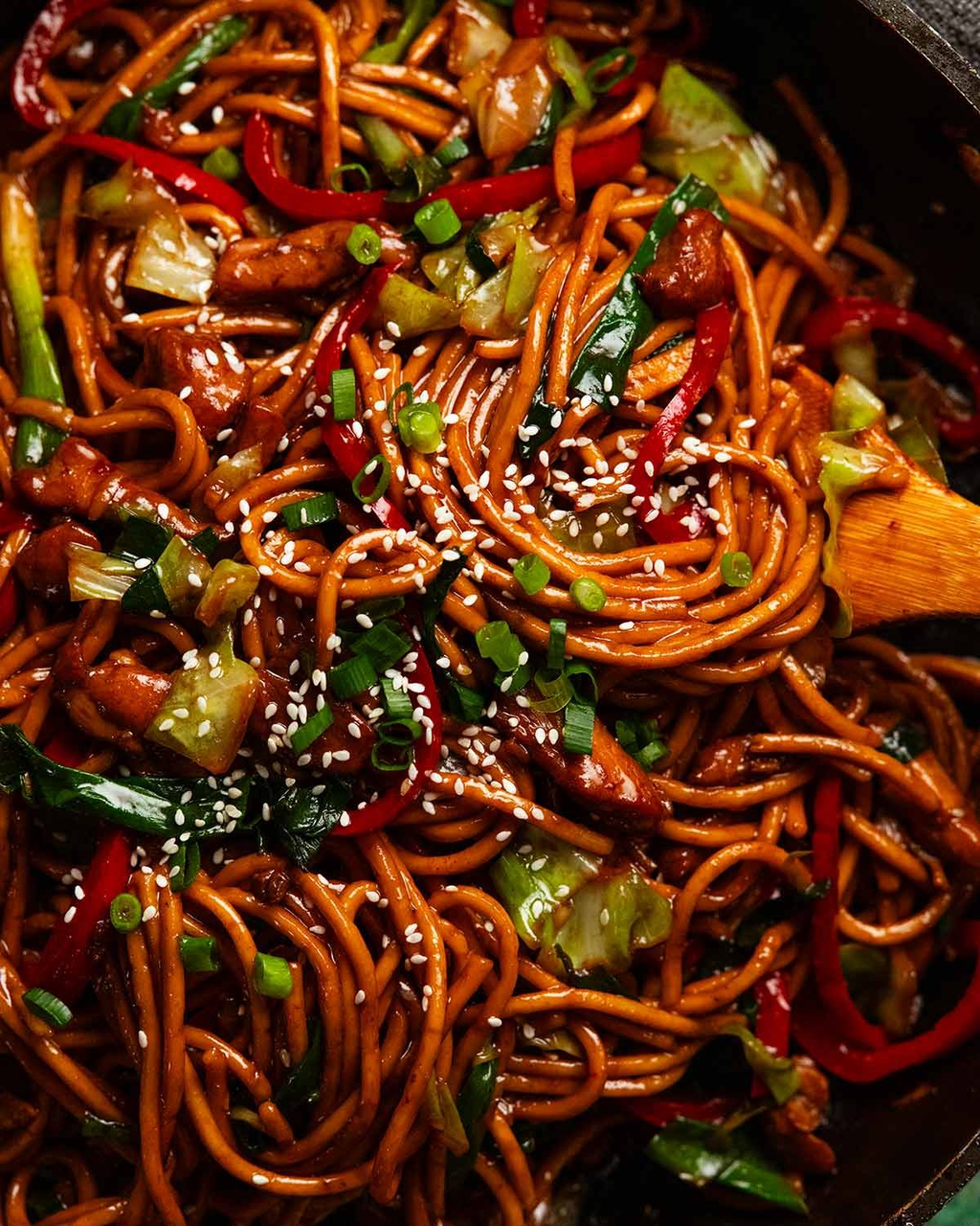 Glossy dark-brown noodles tossed with chicken, red pepper, cabbage and spring onion in a pan, topped with sesame seeds